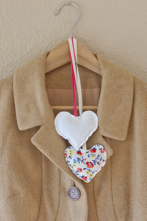 Two heart shaped fabric sachets, one white and one floral, hanging by a ribbon on a camel coat on a wooden hanger